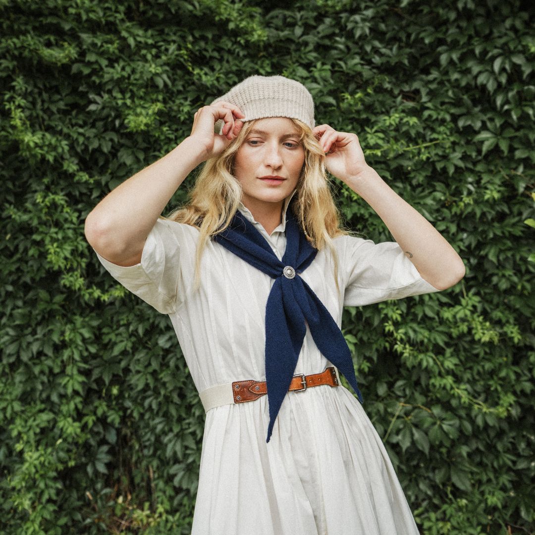 A woman standing outdoors wearing a cream knit beret, a white dress, and a navy blue neck scarf. The background features a lush green ivy-covered wall with dappled sunlight.