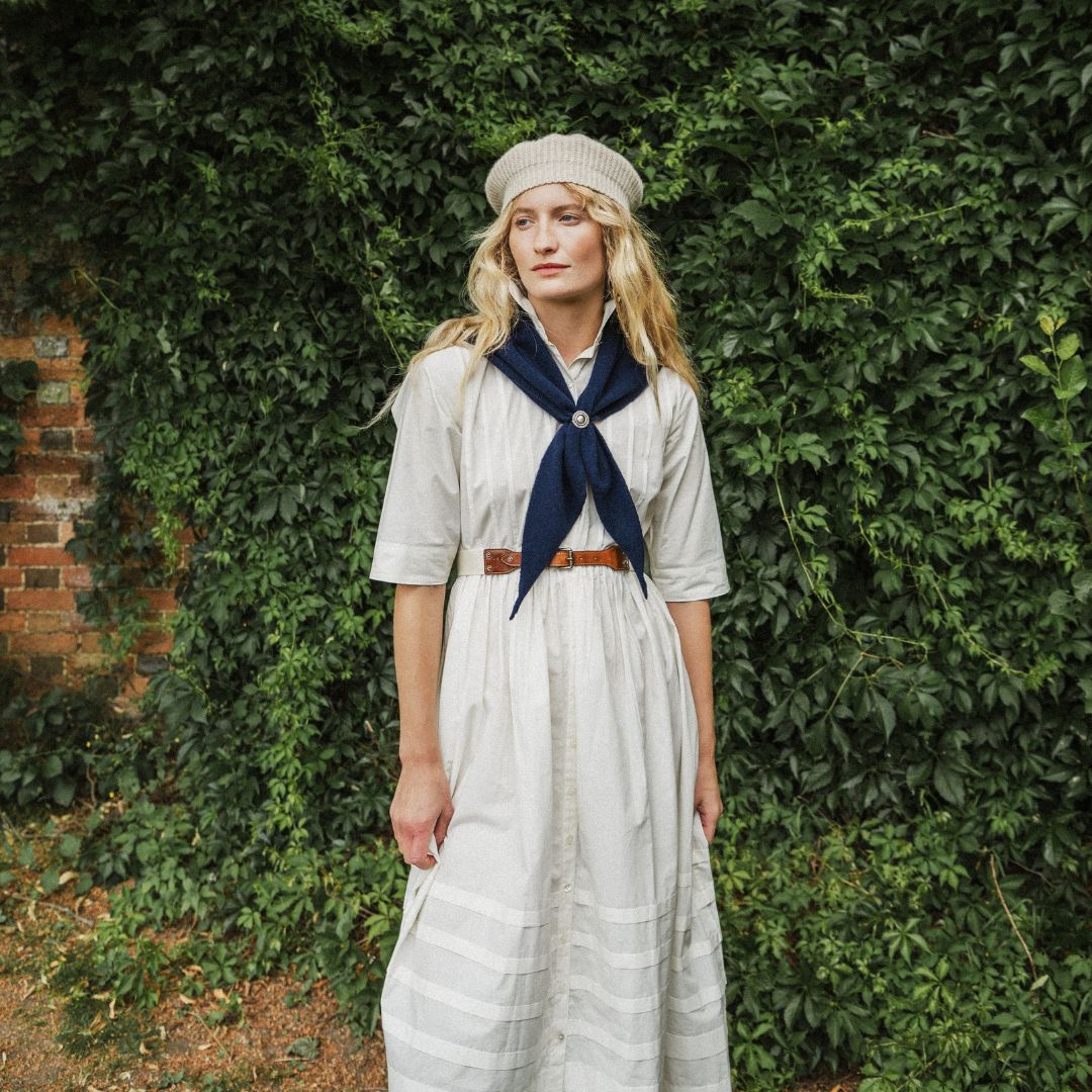 A woman stands outdoors in front of a dense green ivy-covered wall, wearing a long white dress with a navy blue neck scarf, a brown belt, and a beige beret.