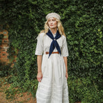 A woman stands outdoors in front of a dense green ivy-covered wall, wearing a long white dress with a navy blue neck scarf, a brown belt, and a beige beret.