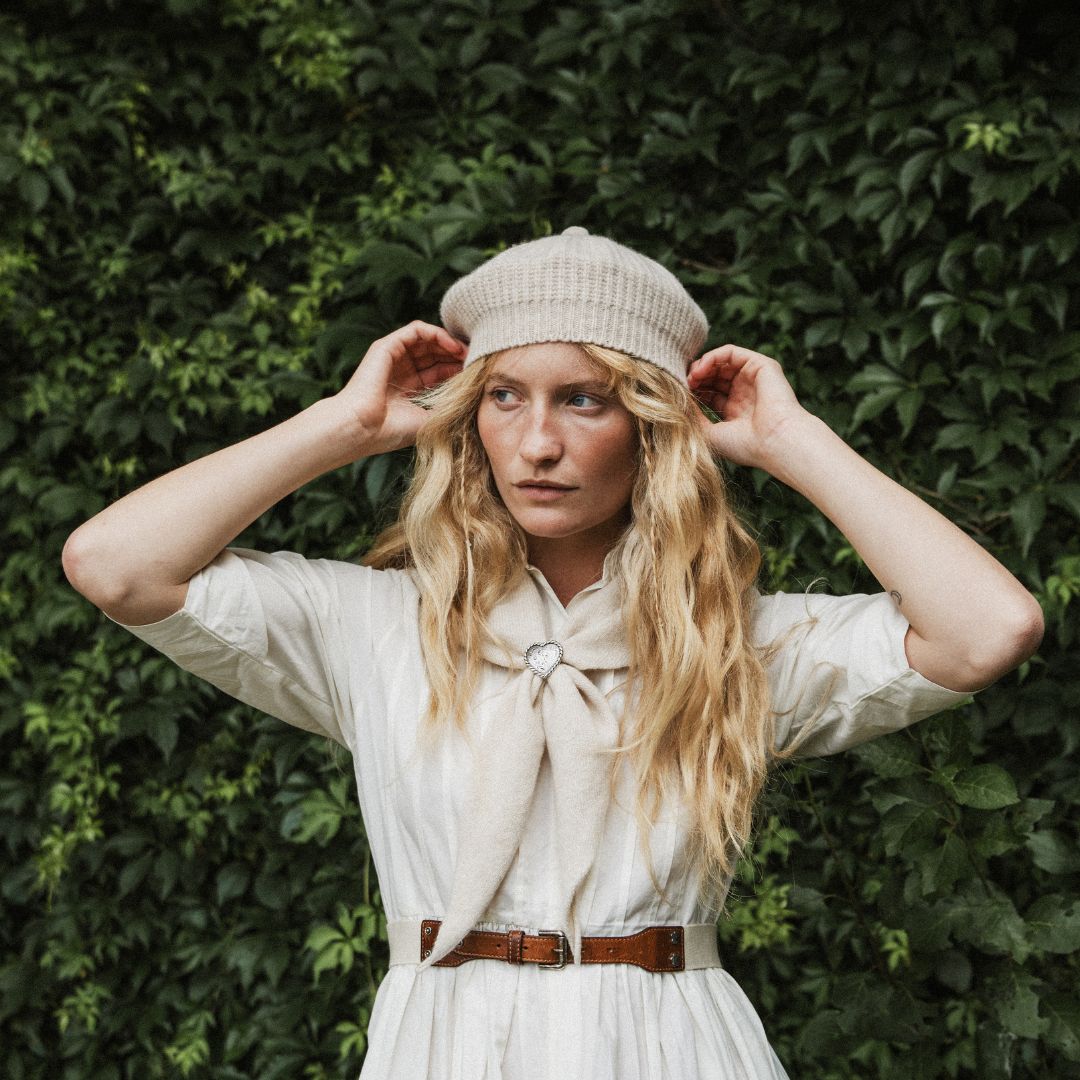 A woman standing outdoors, wearing a cream knit beret, a white dress, and a cream neck scarf. The background features a lush green ivy-covered wall with dappled sunlight.