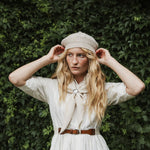 A woman standing outdoors, wearing a cream knit beret, a white dress, and a cream neck scarf. The background features a lush green ivy-covered wall with dappled sunlight.