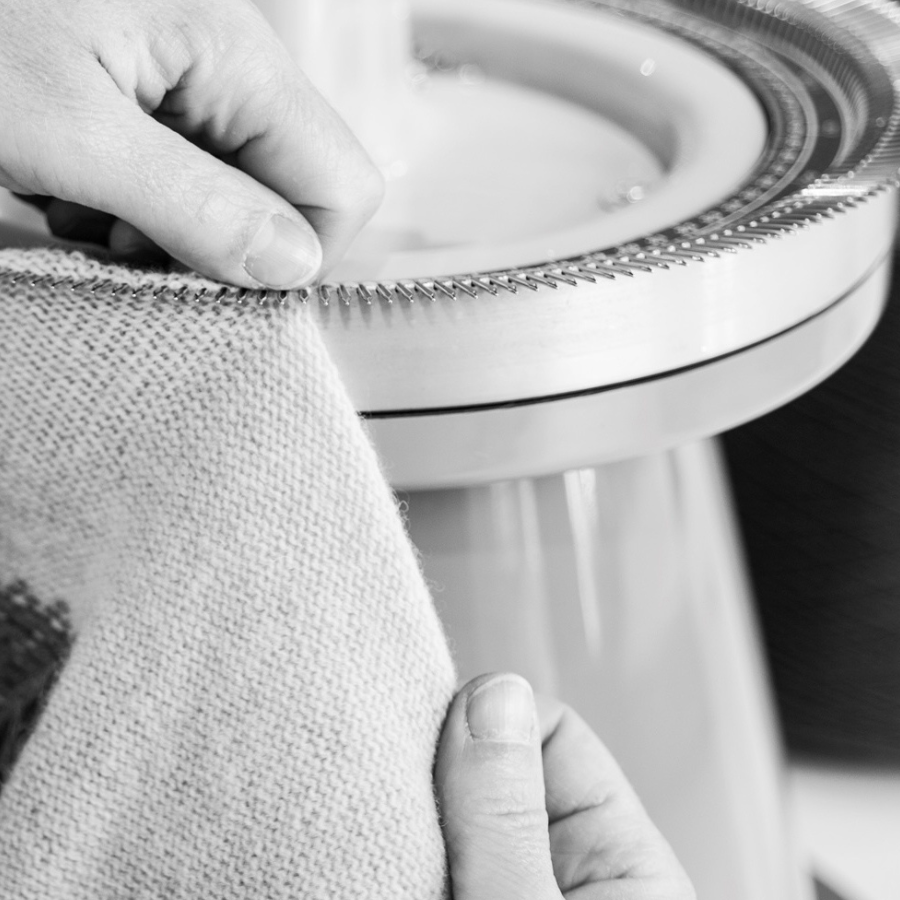 A close-up black and white photo of a person's hands holding a piece of textured fabric next to a circular knitting machine.