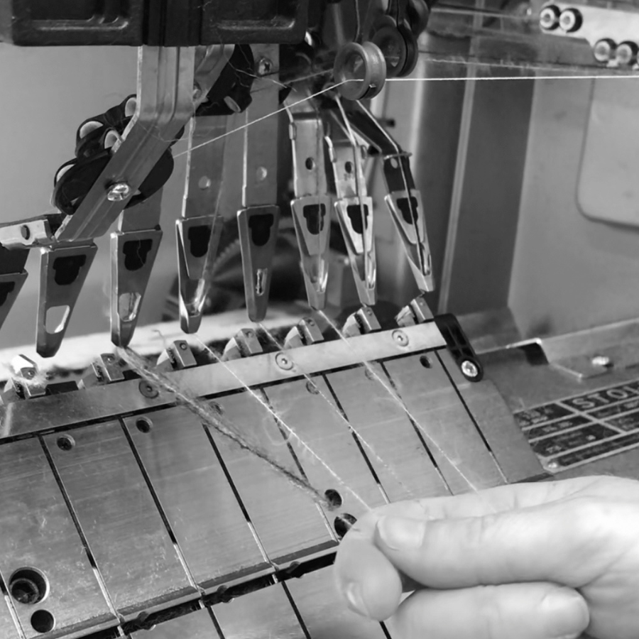 A close-up black and white photo of a hand guiding thread under a knitting machine needle assembly.
