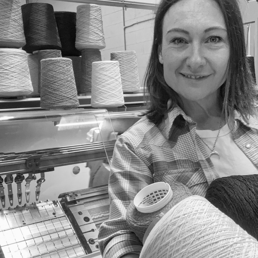 A woman smiling while holding a large spool of yarn in front of a knitting machine with multiple yarn cones on a shelf.