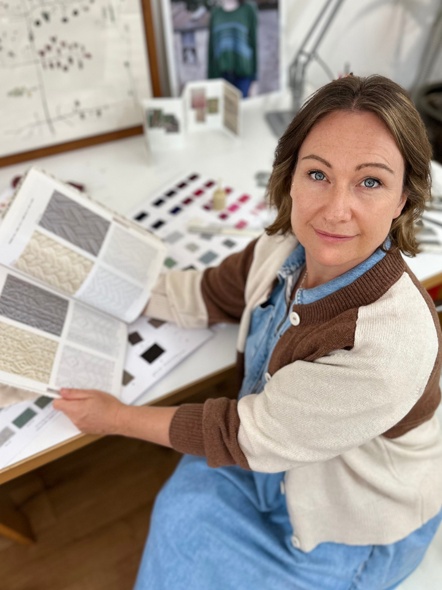 A woman in a home or studio setting, holding an open fabric or wallpaper sample book with various textured swatches. She is wearing a brown and cream cardigan over a blue denim shirt and blue jeans.