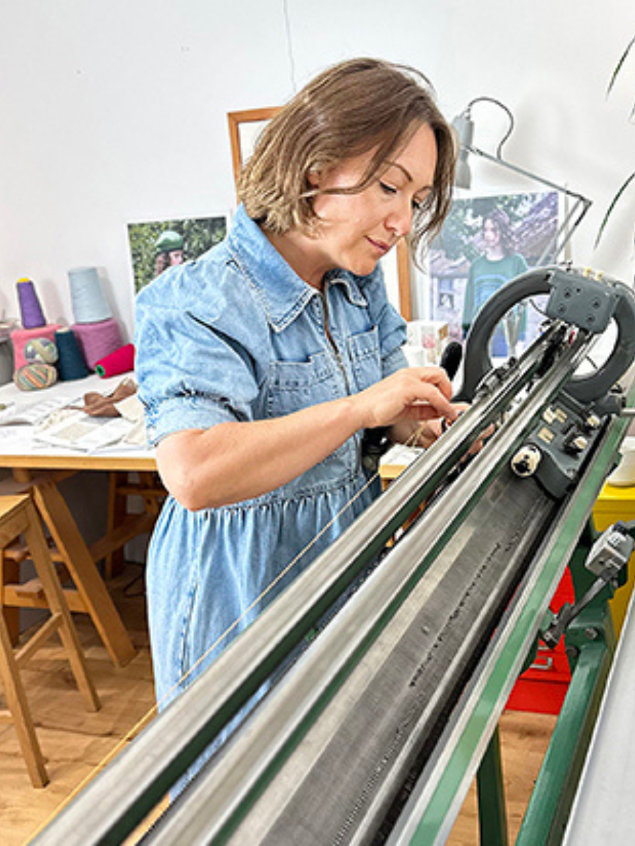 A woman in a light blue denim dress operating a large knitting machine in a bright, creative studio space.