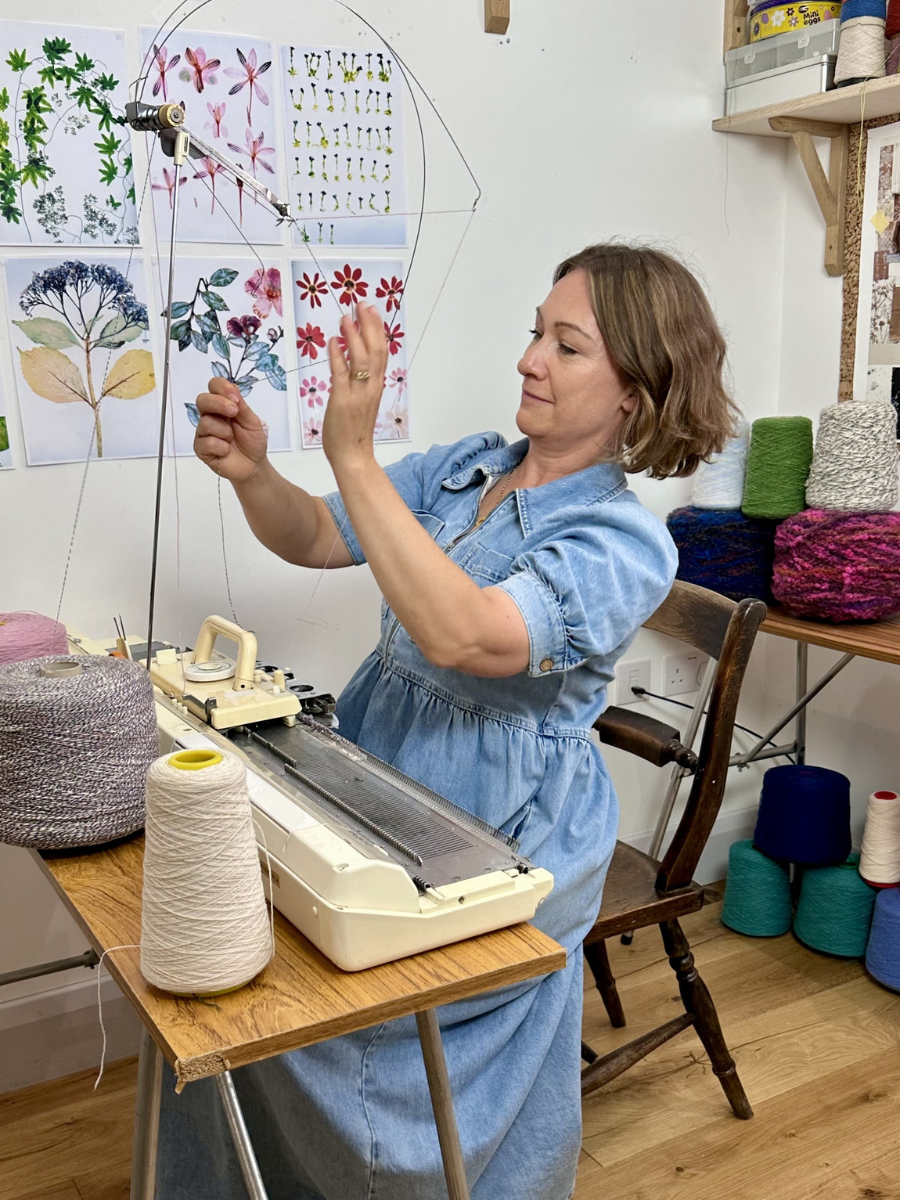 A woman in a blue dress operates a knitting machine in a cozy craft room, with spools of yarn and framed floral artwork visible in the background.