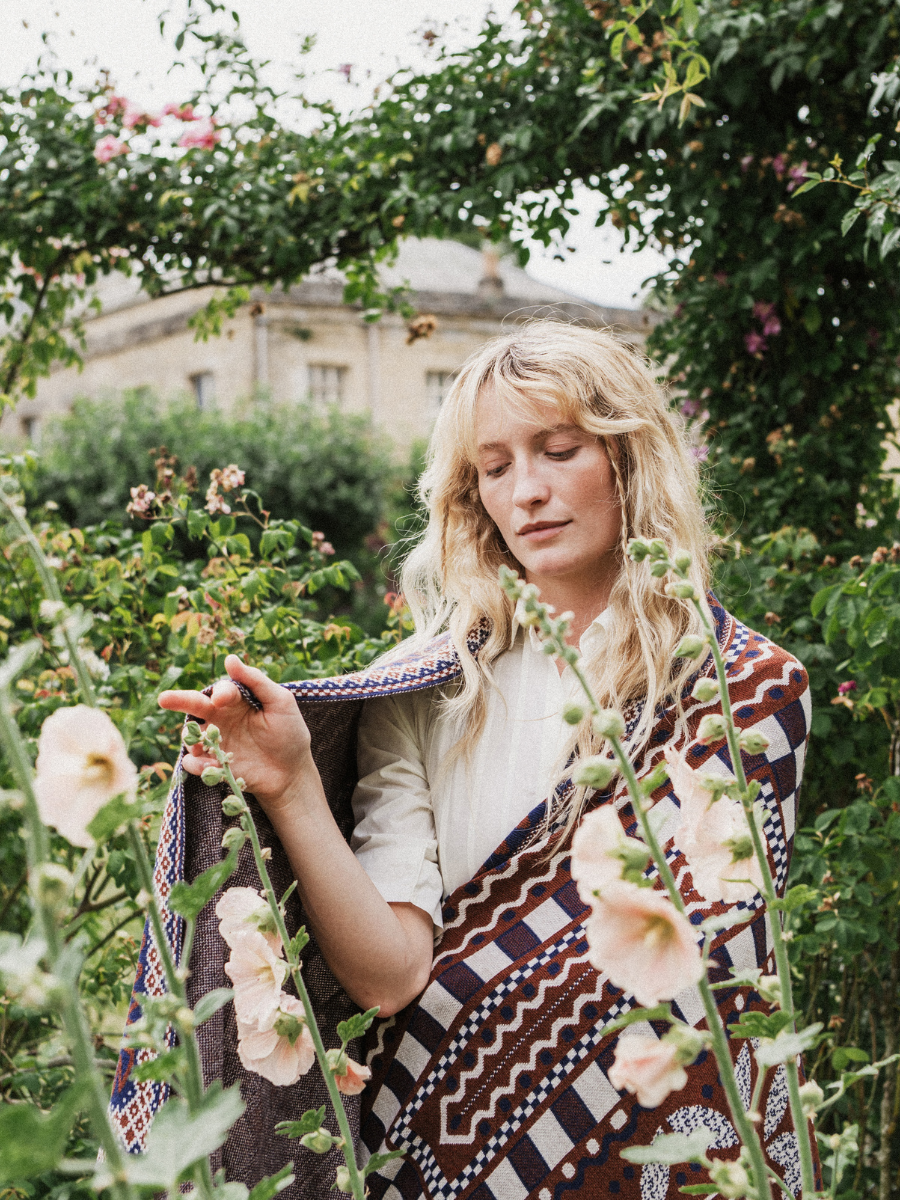 A woman standing in a lush garden, wrapped in a patterned bohemian throw blanket with brown, cream, and purple geometric designs.