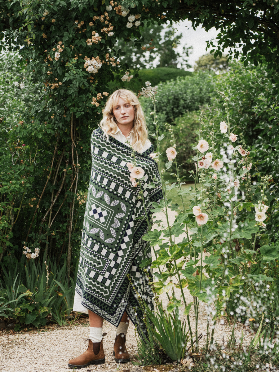 A woman stands in a garden wearing a blanket with a blue, white, and green geometric pattern. She is also wearing brown boots, white socks, and a white dress.