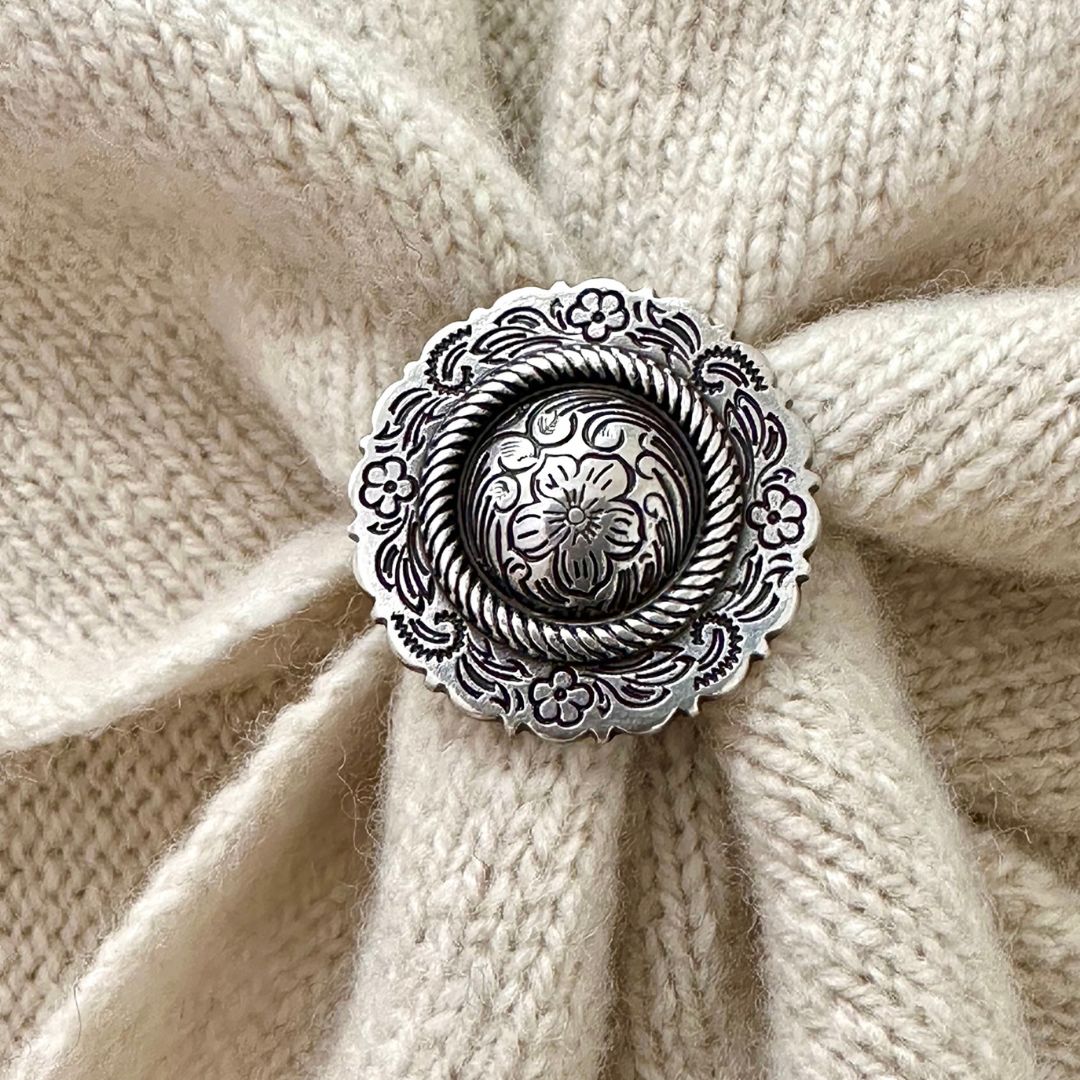 An ornate silver-toned metal scarf slider  with floral engravings and a rope border, placed on a cream-colored knitted fabric background.