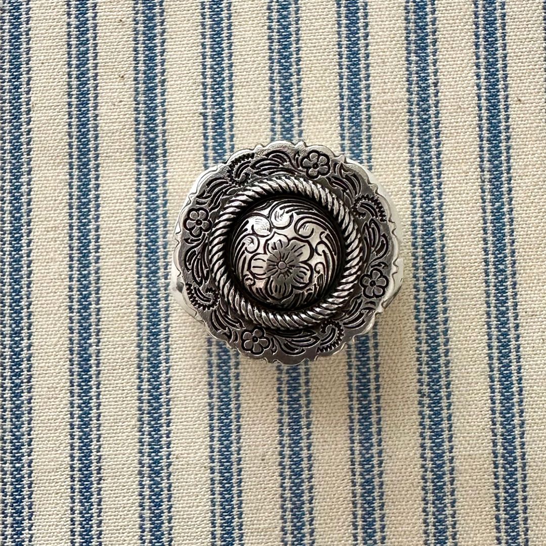 An ornate round silver-toned metal scarf slider with floral and rope-like embossed details, placed on a cream fabric background with blue vertical stripes.