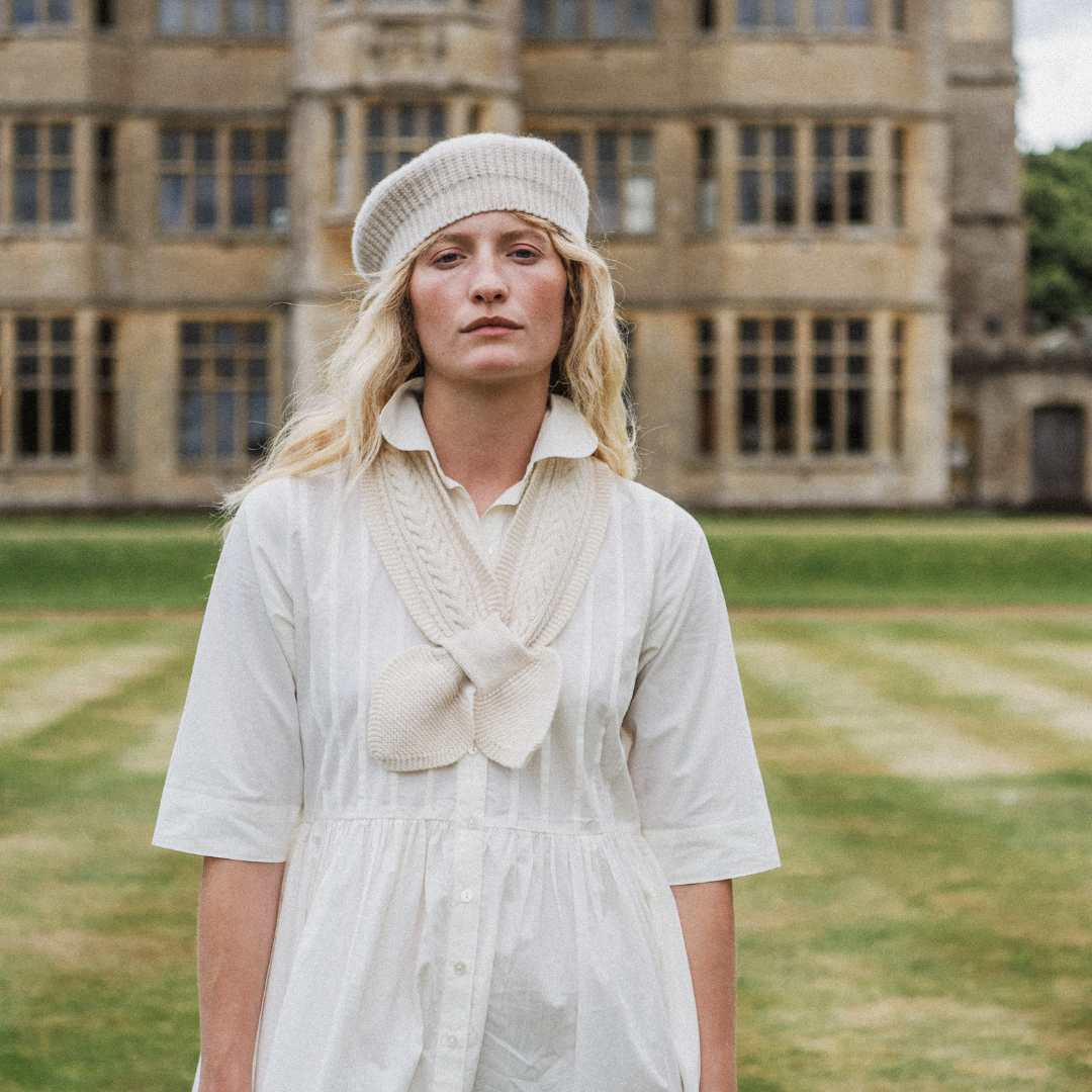 A woman stands outdoors in front of a large stone building on a grassy lawn, wearing a cream-colored knit beret and a matching cream knit bow detail over a white short-sleeve dress.
