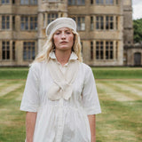 A woman stands outdoors in front of a large stone building on a grassy lawn, wearing a cream-colored knit beret and a matching cream knit bow detail over a white short-sleeve dress.