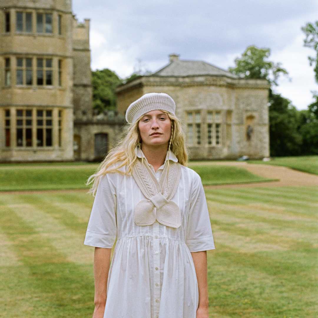 A woman stands on a manicured lawn in front of a large stone mansion, wearing a white dress with a knitted scarf at the  neckline and a matching white knit hat.