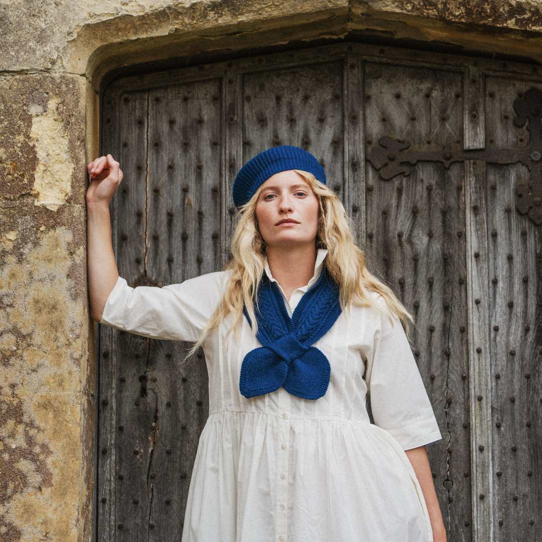 A woman stands in front of a rustic wooden door wearing a cream-colored dress, a blue knit beret, and a matching blue knotted scarf.