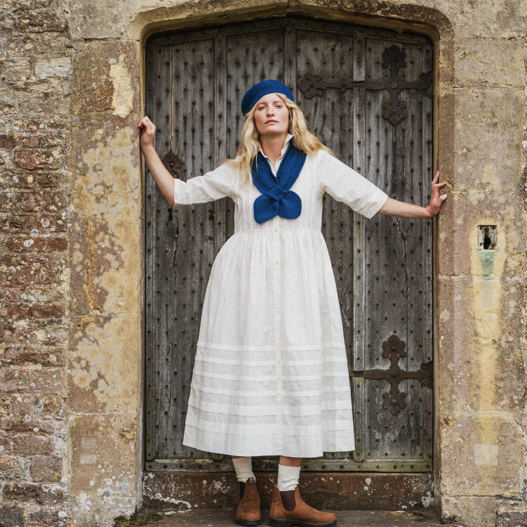 A woman stands in front of an old stone wall and weathered wooden door, wearing a white midi dress with a long blue scarf at the neckline, a blue headband, white socks, and brown boots.