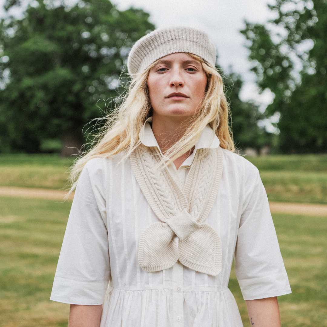 A woman standing outdoors in a grassy field wearing a cream-colored knit beanie, a cream knit collar detail over a white dress, and a white shirt. Trees are blurred in the background.