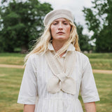 A woman standing outdoors in a grassy field wearing a cream-colored knit beanie, a cream knit collar detail over a white dress, and a white shirt. Trees are blurred in the background.