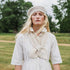A woman standing outdoors in a grassy field wearing a cream-colored knit beanie, a cream knit collar detail over a white dress, and a white shirt. Trees are blurred in the background.