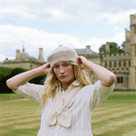 A woman wearing a cream-colored knit beanie and matching cream blouse stands in a grassy field with a large stone castle in the background, adjusting her beanie.
