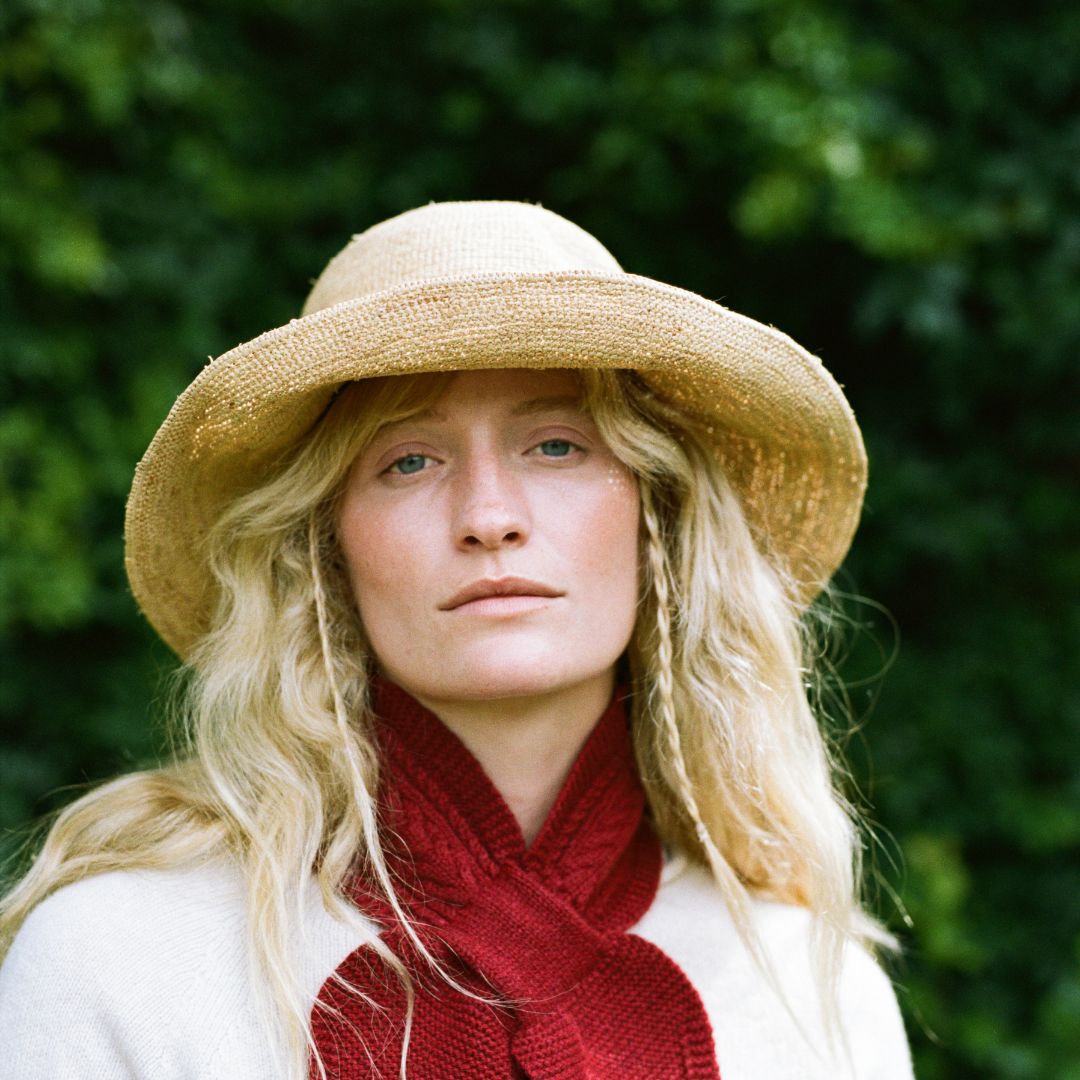 A woman outdoors wearing a wide-brimmed straw hat, a deep red knit scarf, and a cream-colored top, with a soft-focus green foliage background.