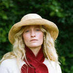 A woman outdoors wearing a wide-brimmed straw hat, a deep red knit scarf, and a cream-colored top, with a soft-focus green foliage background.