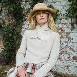 A woman wearing a cream knit sweater with a large bow collar, a straw hat, and a plaid skirt, sitting against a weathered white brick wall with green ivy.