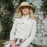 A woman wearing a cream knit sweater with a large bow collar, a straw hat, and a plaid skirt, sitting against a weathered white brick wall with green ivy.