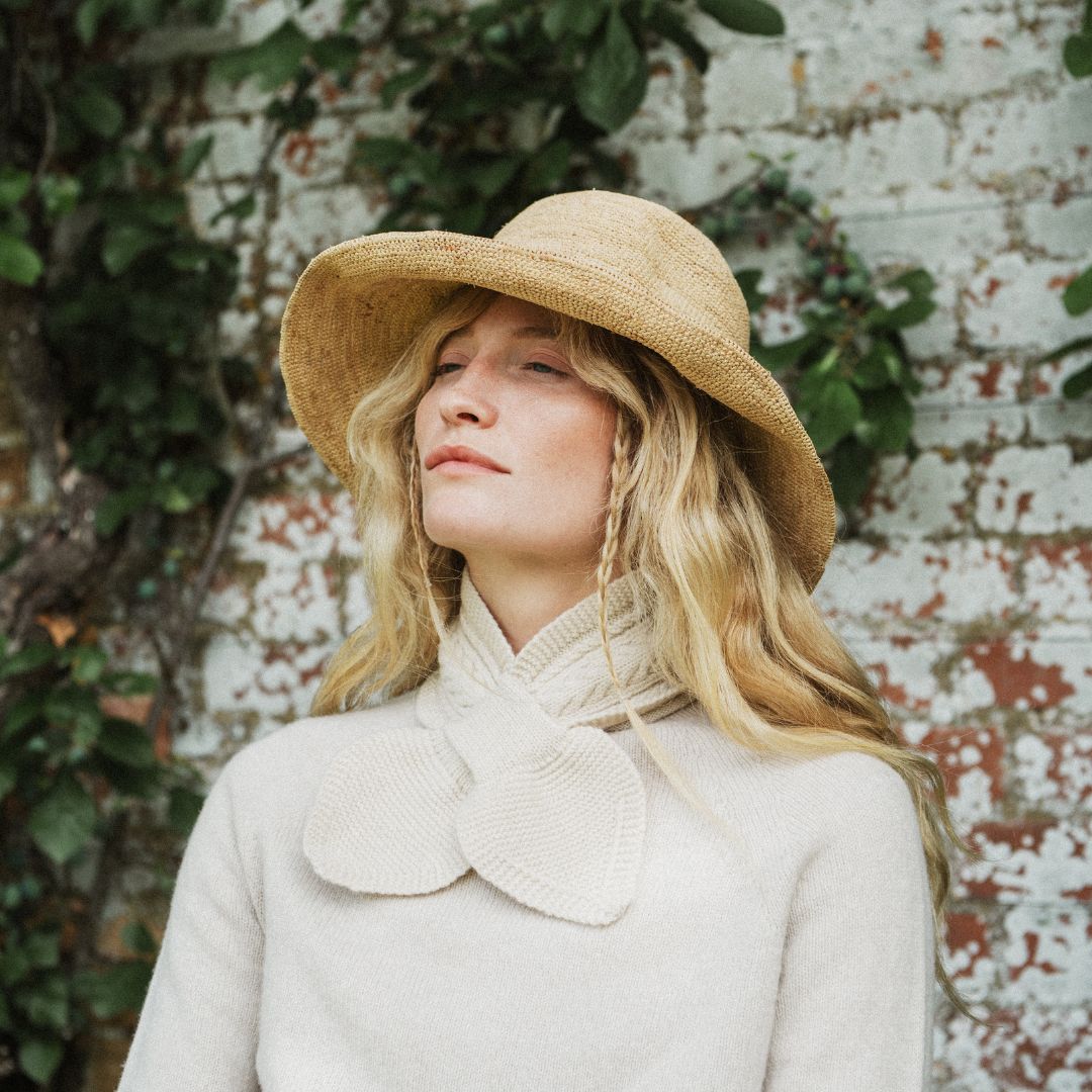 A woman wearing a wide-brimmed straw hat and a cream knit sweater with a large bow collar, standing against a white brick wall with green ivy.