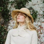 A woman wearing a wide-brimmed straw hat and a cream knit sweater with a large bow collar, standing against a white brick wall with green ivy.