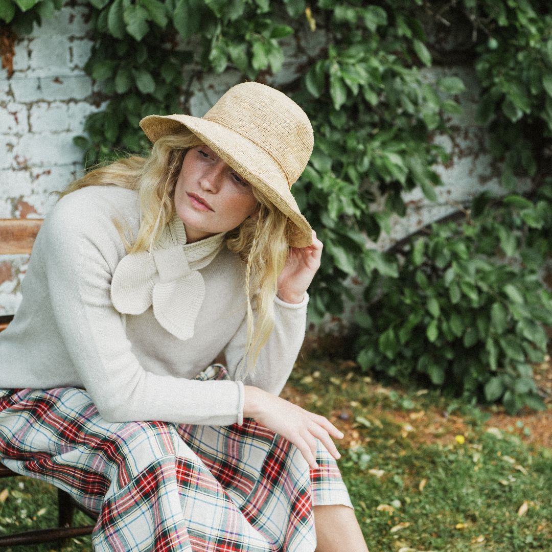 A woman sitting outdoors on a bench in front of a white brick wall covered in green ivy. She is wearing a wide-brimmed straw hat, a cream knit sweater with a large bow detail at the neck, and a red, green, and white plaid skirt.