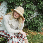 A woman sitting outdoors on a bench in front of a white brick wall covered in green ivy. She is wearing a wide-brimmed straw hat, a cream knit sweater with a large bow detail at the neck, and a red, green, and white plaid skirt.