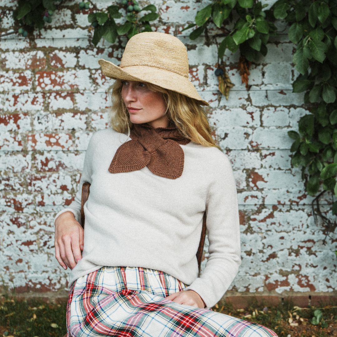 A woman wearing a cream knit sweater, a brown knit bow collar, a straw hat, and a red, green, and white plaid skirt, sitting in front of a weathered white brick wall with green ivy.