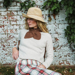 A woman wearing a cream knit sweater, a brown knit bow collar, a straw hat, and a red, green, and white plaid skirt, sitting in front of a weathered white brick wall with green ivy.