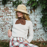 A woman wearing a cream knit sweater, a brown knit bow collar, a straw hat, and a red, green, and white plaid skirt, sitting in front of a weathered white brick wall with green ivy.