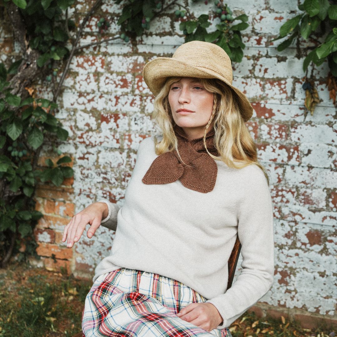A woman wearing a cream knit sweater with a brown knit bow collar, a straw hat, and a red, green, and white plaid skirt, posing in front of a weathered white brick wall with green vines.