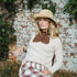 A woman wearing a cream knit sweater with a brown knit bow collar, a straw hat, and a red, green, and white plaid skirt, posing in front of a weathered white brick wall with green vines.
