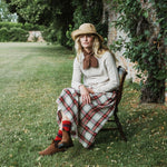 A woman sits on a wooden chair in a grassy garden setting, wearing a wide-brimmed straw hat, a cream knit sweater, a brown bow tie, a red and green plaid skirt, striped socks, and brown boots.