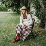 A woman sits on a wooden chair in a grassy garden setting, wearing a wide-brimmed straw hat, a cream knit sweater, a brown bow tie, a red and green plaid skirt, striped socks, and brown boots.
