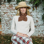 A woman wearing a beige straw hat, a cream knit sweater, a brown knotted scarf, and a red and blue plaid skirt, sitting in front of a weathered white brick wall.