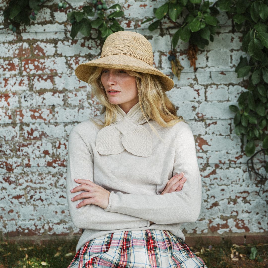 A woman wearing a beige straw hat and a cream-colored knit sweater with a large decorative collar, paired with a red, green, and white plaid skirt, posing in front of a weathered white brick wall with green ivy.
