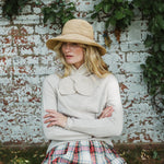 A woman wearing a beige straw hat and a cream-colored knit sweater with a large decorative collar, paired with a red, green, and white plaid skirt, posing in front of a weathered white brick wall with green ivy.