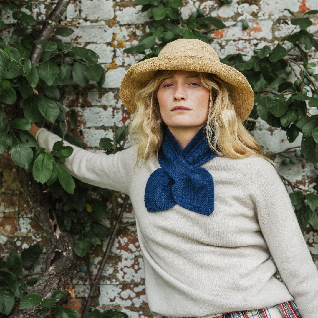 A woman wearing a beige knit sweater, a large navy blue bow tie scarf, and a straw sun hat, posing outdoors against a weathered brick wall with green ivy.