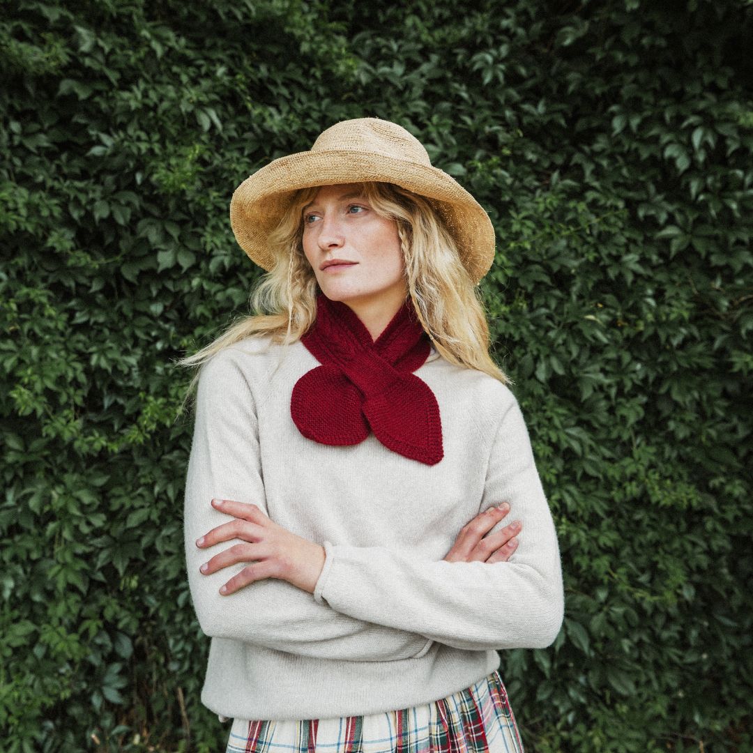 A woman wearing a beige straw hat, a cream knit sweater, and a deep red knitted bow scarf, standing with arms crossed against a dense green ivy wall.