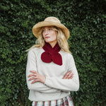 A woman wearing a beige straw hat, a cream knit sweater, and a deep red knitted bow scarf, standing with arms crossed against a dense green ivy wall.