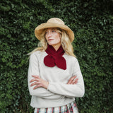 A woman wearing a beige straw hat, a cream knit sweater, and a deep red knitted bow scarf, standing with arms crossed against a dense green ivy wall.