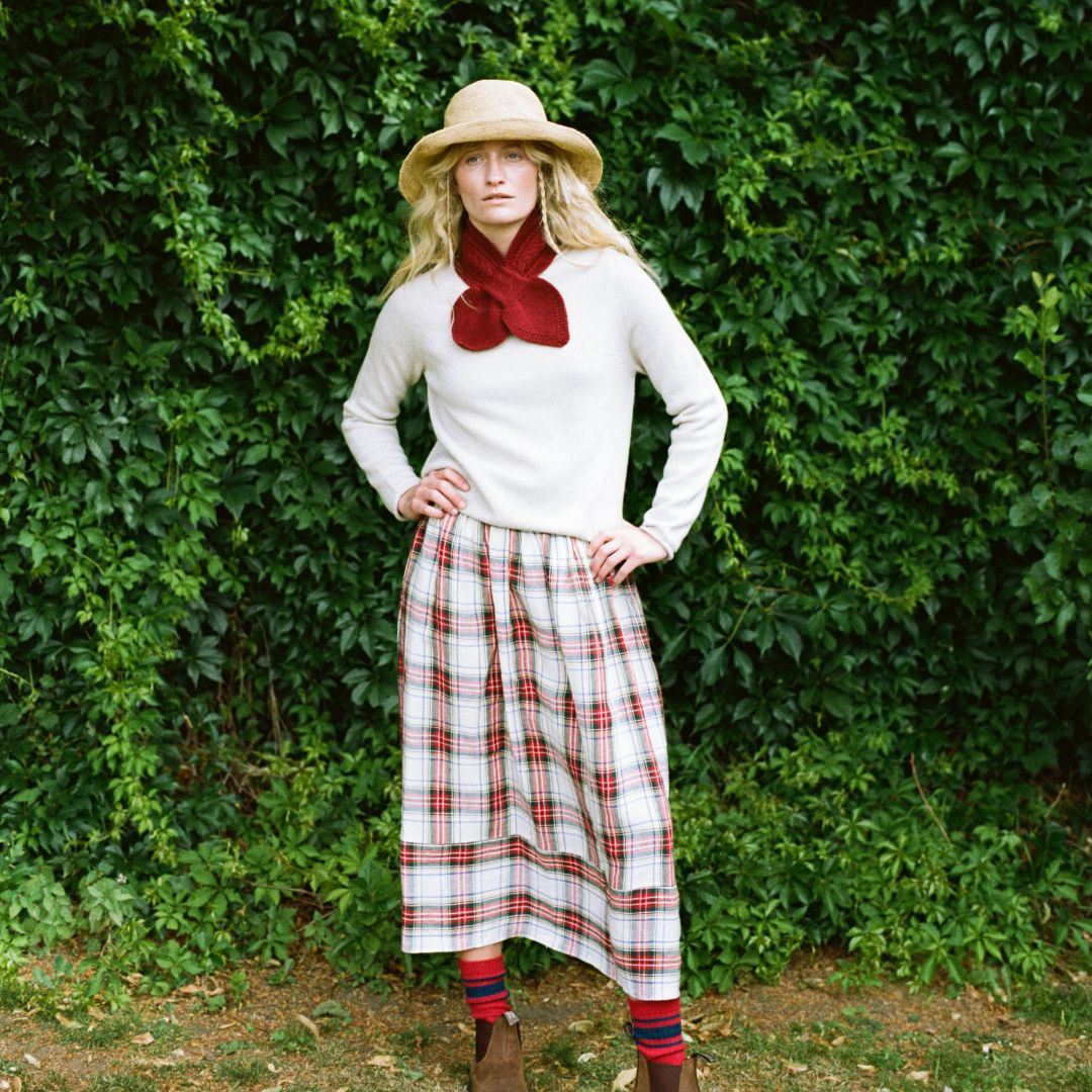 A woman stands outdoors in front of a dense green hedge, wearing a cream knit sweater, a large dark red bow tie, a red and white plaid skirt, a straw hat, and brown boots.