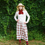 A woman stands outdoors in front of a dense green hedge, wearing a cream knit sweater, a large dark red bow tie, a red and white plaid skirt, a straw hat, and brown boots.