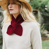 A woman wearing a cream knit sweater, a straw hat, and a deep burgundy knitted bow scarf around her neck, photographed outdoors with a blurred garden background.