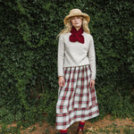 A woman stands outdoors in front of a dense green hedge, wearing a cream knit sweater, a red bow at the collar, a red and green plaid midi skirt, a straw hat, and red socks.